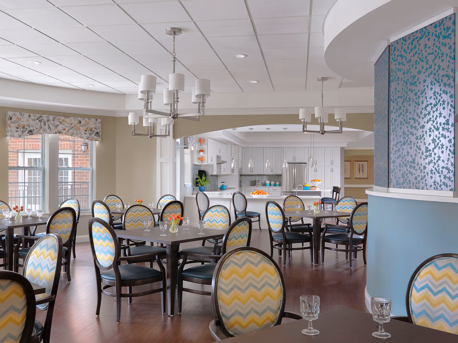 A bright dining room with multiple dark wood tables and chairs featuring chevron-patterned upholstery in yellow, blue, and white. The room has large windows with floral valances, modern chandeliers, and a view into a white kitchen with stainless steel appliances and a countertop with bowls of oranges.