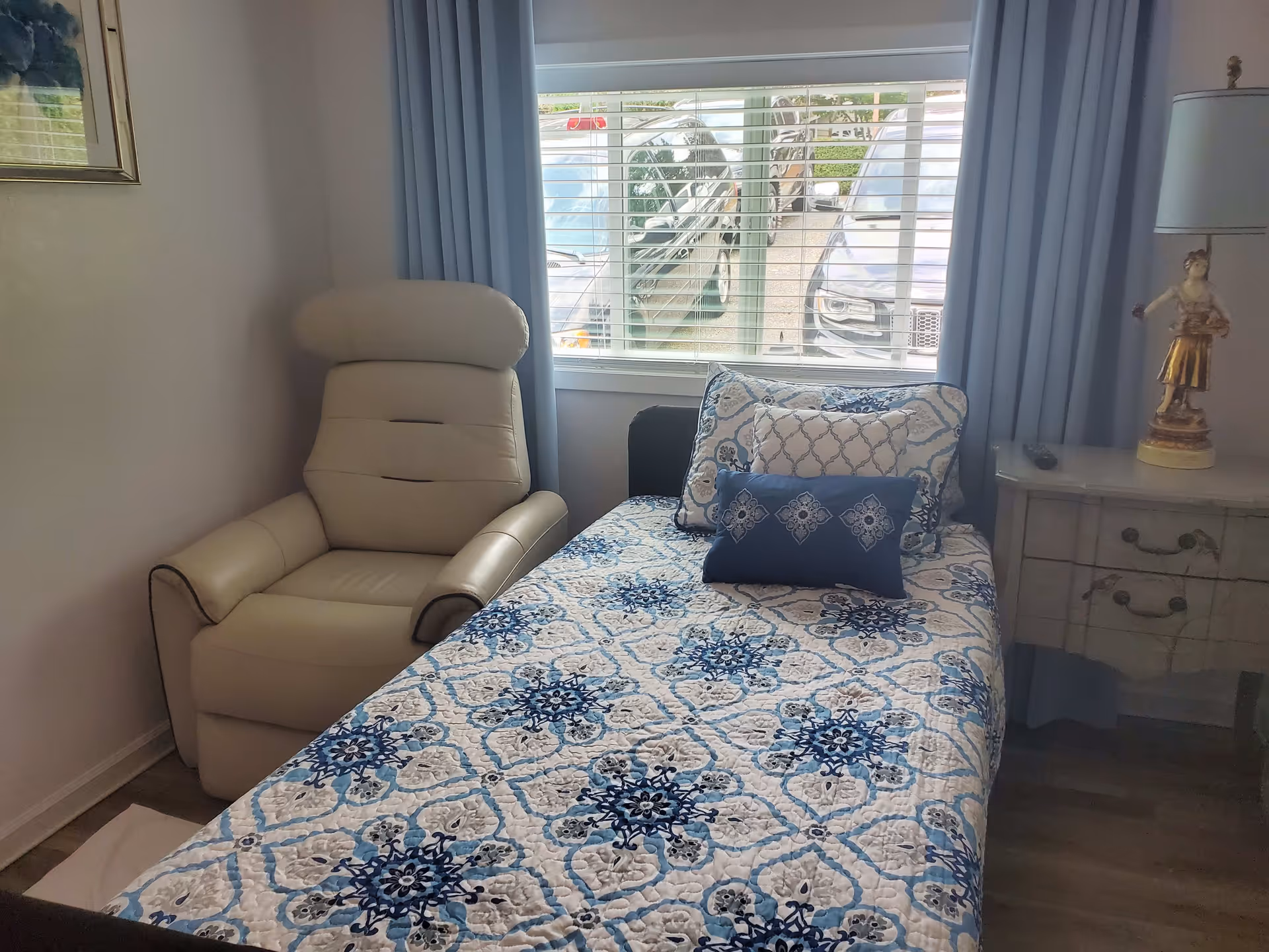 Small bedroom with a single bed covered in a blue patterned quilt, a beige recliner, nightstand and window with blinds showing parked cars outside.
