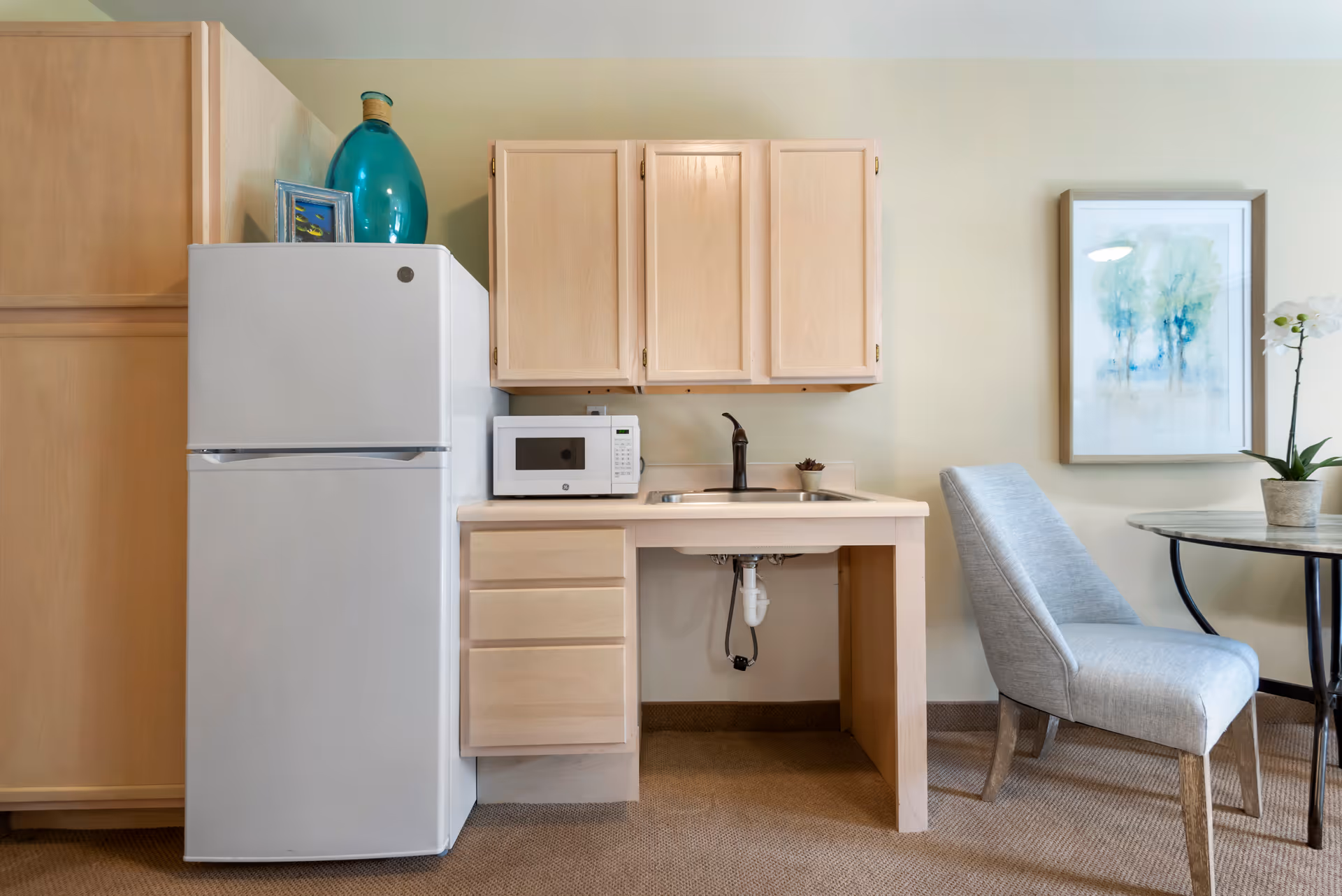 A small kitchenette area with light wood cabinets, a white refrigerator, a white microwave on the counter, and a sink with a black faucet. To the right, there is a light gray upholstered chair next to a round table with a potted orchid and a framed watercolor painting of trees on the wall.