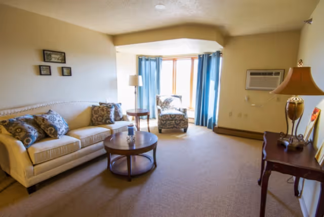 A cozy living room with a beige sofa adorned with patterned cushions, a round wooden coffee table, a patterned armchair near a window with blue curtains, a floor lamp, an air conditioning unit mounted on the wall, and a wooden side table with a decorative lamp and framed pictures.