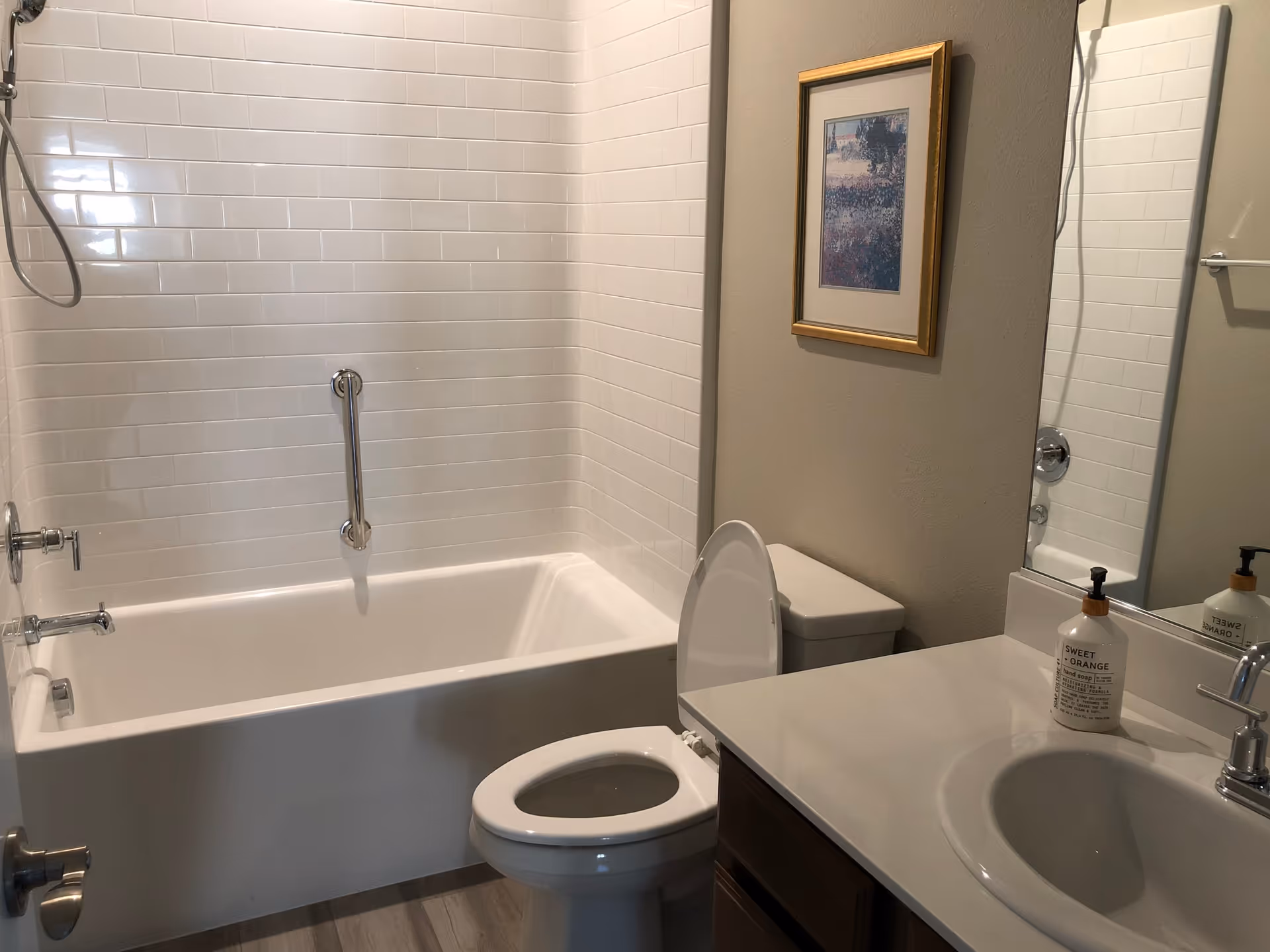 A clean bathroom featuring a white bathtub with a showerhead and grab bar, a toilet with the lid open, a sink with a soap dispenser labeled 'Sweet Orange,' a large mirror, and a framed picture on the wall.