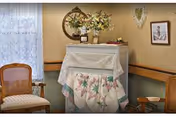 A cozy corner of a room featuring two wooden chairs with cushioned seats, a small table covered with a floral quilted cloth, a round mirror on the wall above the table, a flower arrangement in a vase, and framed artwork on the wall. The room has beige walls and a window with lace curtains.