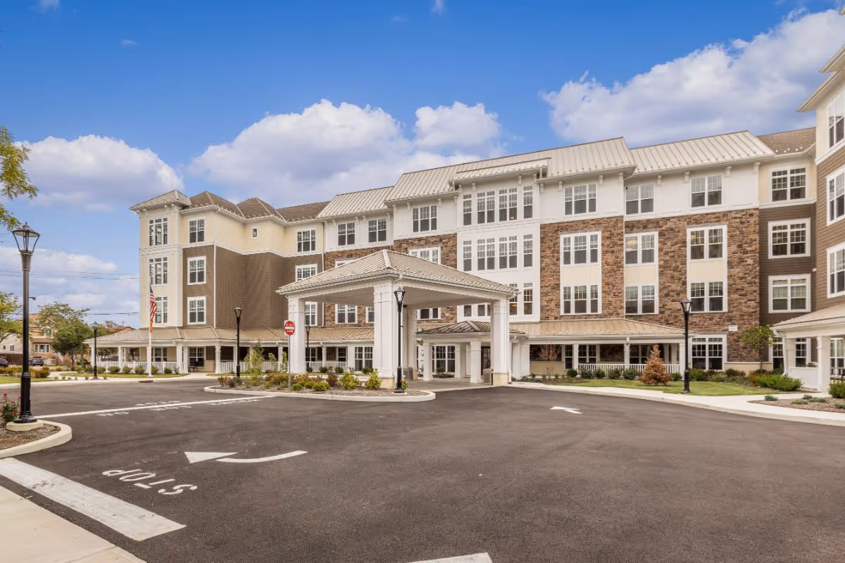 Front entrance of a multi-story senior living facility with a covered porte-cochère, landscaped driveway, and an American flag.