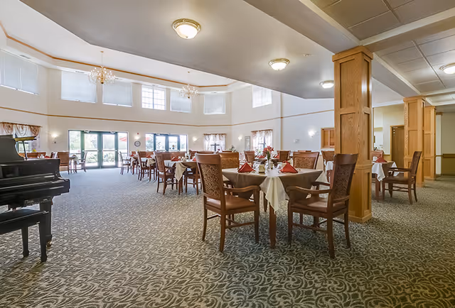 Spacious dining room with multiple tables covered in white tablecloths and set with red napkins and tableware. The room features large windows allowing natural light, a patterned carpet, wooden columns, and a black grand piano on the left side.