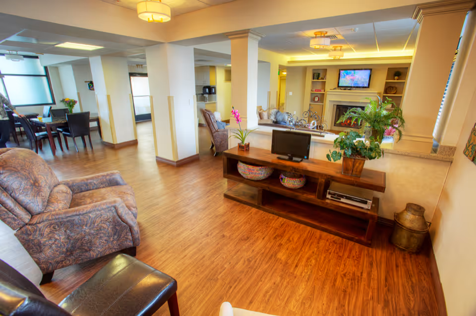 A bright and spacious common area in a senior living facility featuring wooden flooring, a patterned armchair, a wooden console table with plants and a small TV, and a fireplace with a mounted TV above it. In the background, there is a dining area with tables and chairs and large windows allowing natural light to enter.