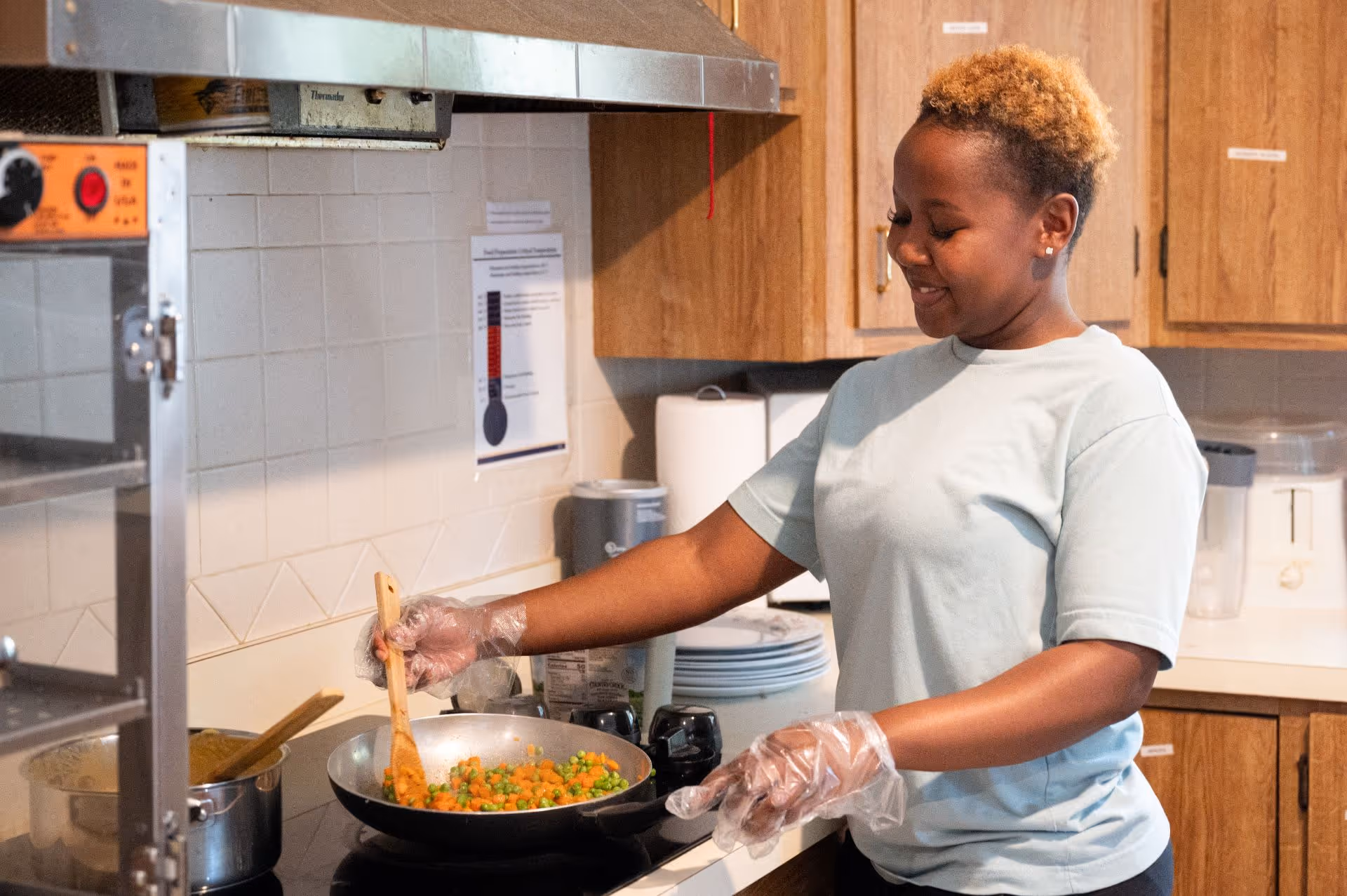 A person wearing a light blue shirt and disposable gloves is cooking mixed vegetables in a frying pan on a stove in a kitchen with wooden cabinets and white tiled walls.