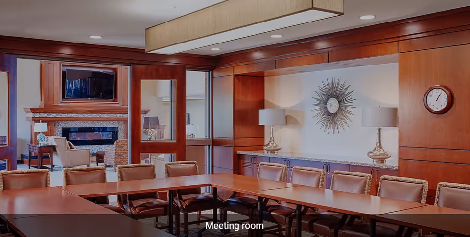 A meeting room with a large U-shaped wooden table surrounded by brown leather chairs. The room features wood-paneled walls, a decorative sunburst wall art, two table lamps on a sideboard, and a clock on the wall. Through glass doors, a cozy lounge area with armchairs, a fireplace, and a mounted TV is visible.