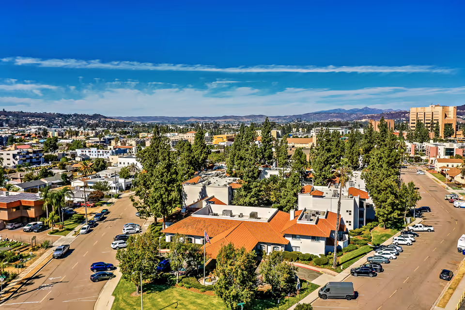 Aerial exterior view of a senior living complex with red-tiled roofs, trees, parked cars and a city skyline with mountains in the distance.