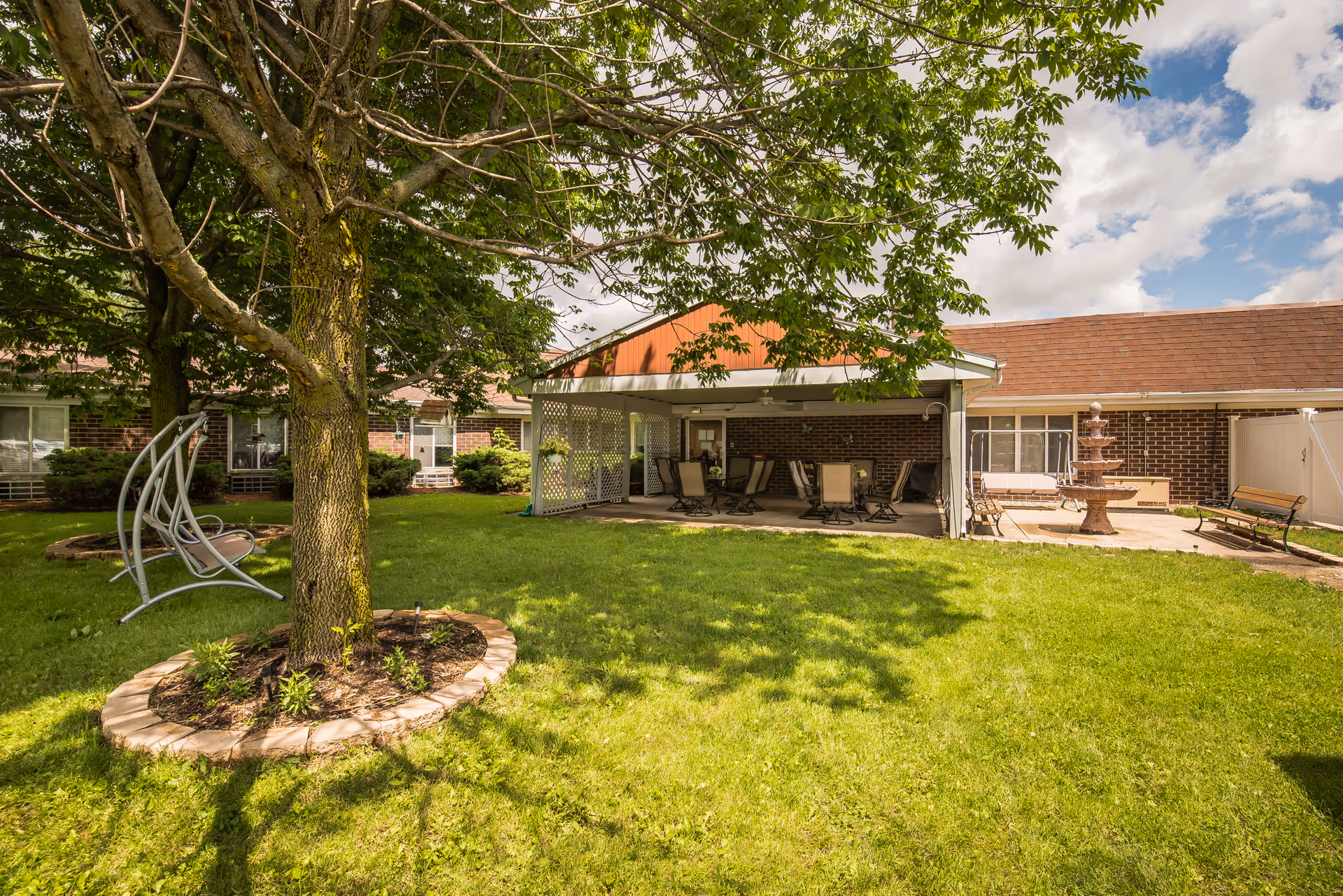 A sunny outdoor garden area at Goldwater Care Spring Valley featuring a large tree with a circular brick border, a metal swing bench, a covered patio with several chairs and tables, a multi-tiered water fountain, and a bench. The building with brick walls and windows surrounds the garden area.