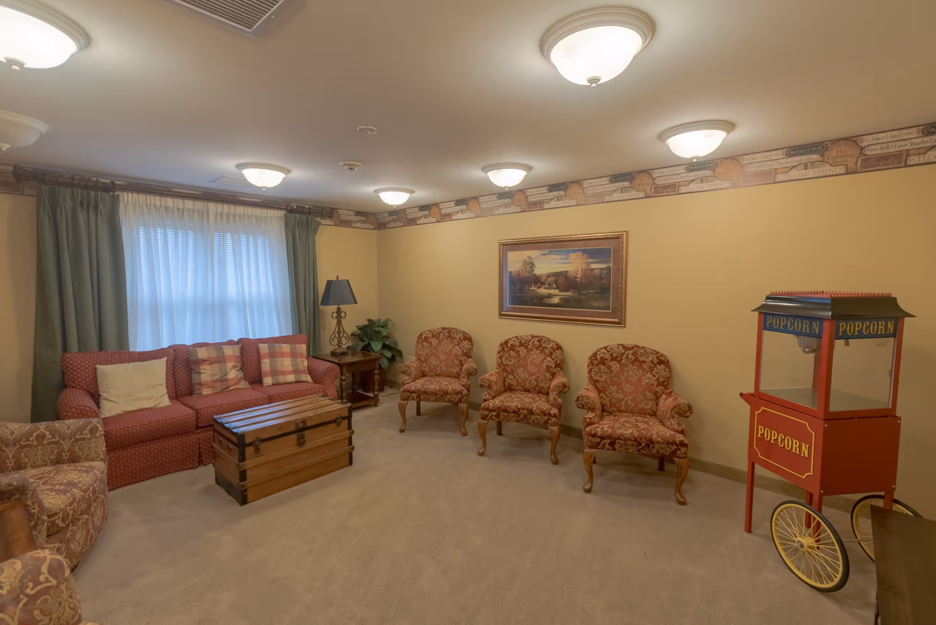 A cozy living room with a red patterned sofa and three matching armchairs arranged along the walls. There is a wooden chest serving as a coffee table in the center, a side table with a lamp and a plant next to the sofa, and a vintage-style red popcorn machine on wheels in the corner. The walls are painted yellow with a decorative border near the ceiling, and a framed landscape painting hangs on the wall above the armchairs. The room is softly lit by multiple ceiling lights.