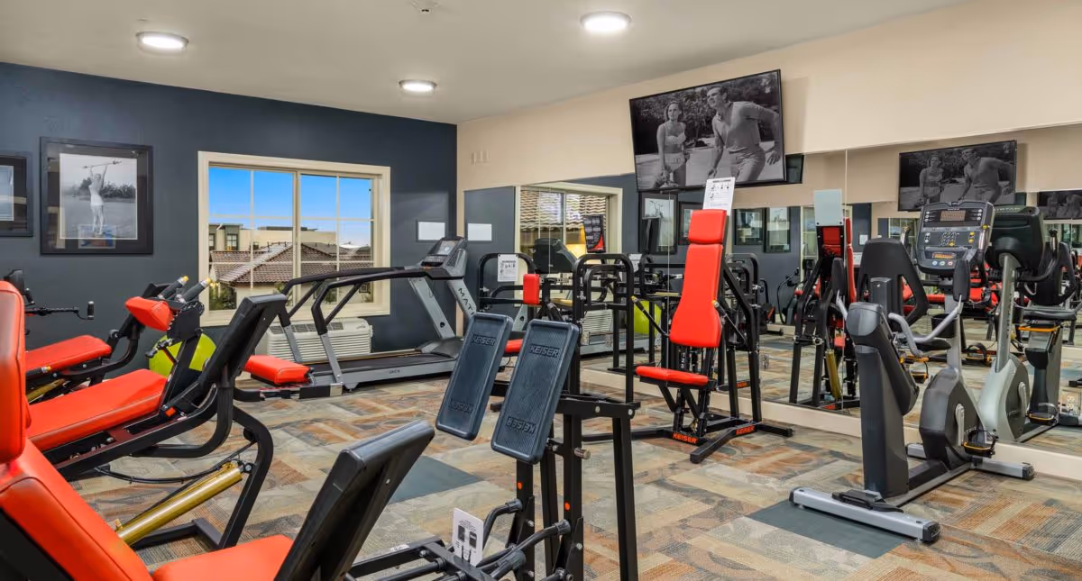Indoor fitness room with treadmills, cardio machines, and red-padded strength equipment reflected in wall mirrors.
