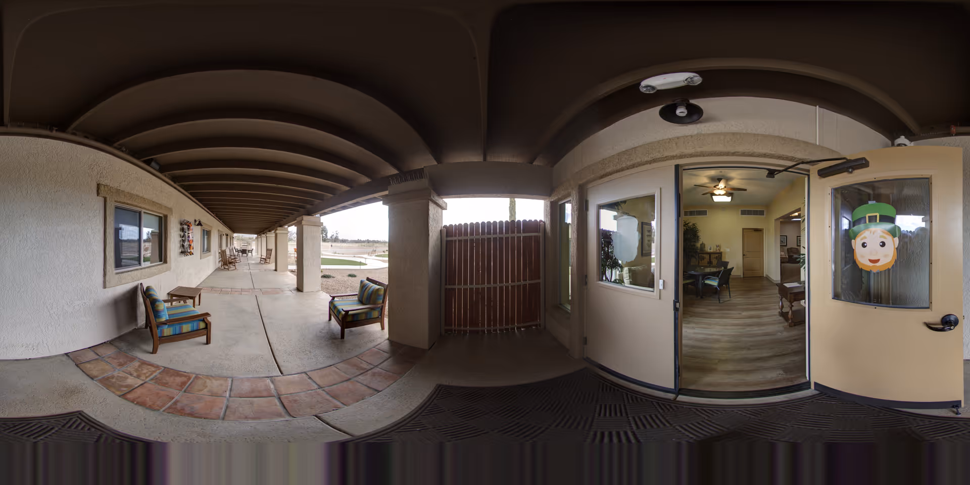 Covered exterior walkway with chairs leading to an open entrance into a common room at an assisted living facility.