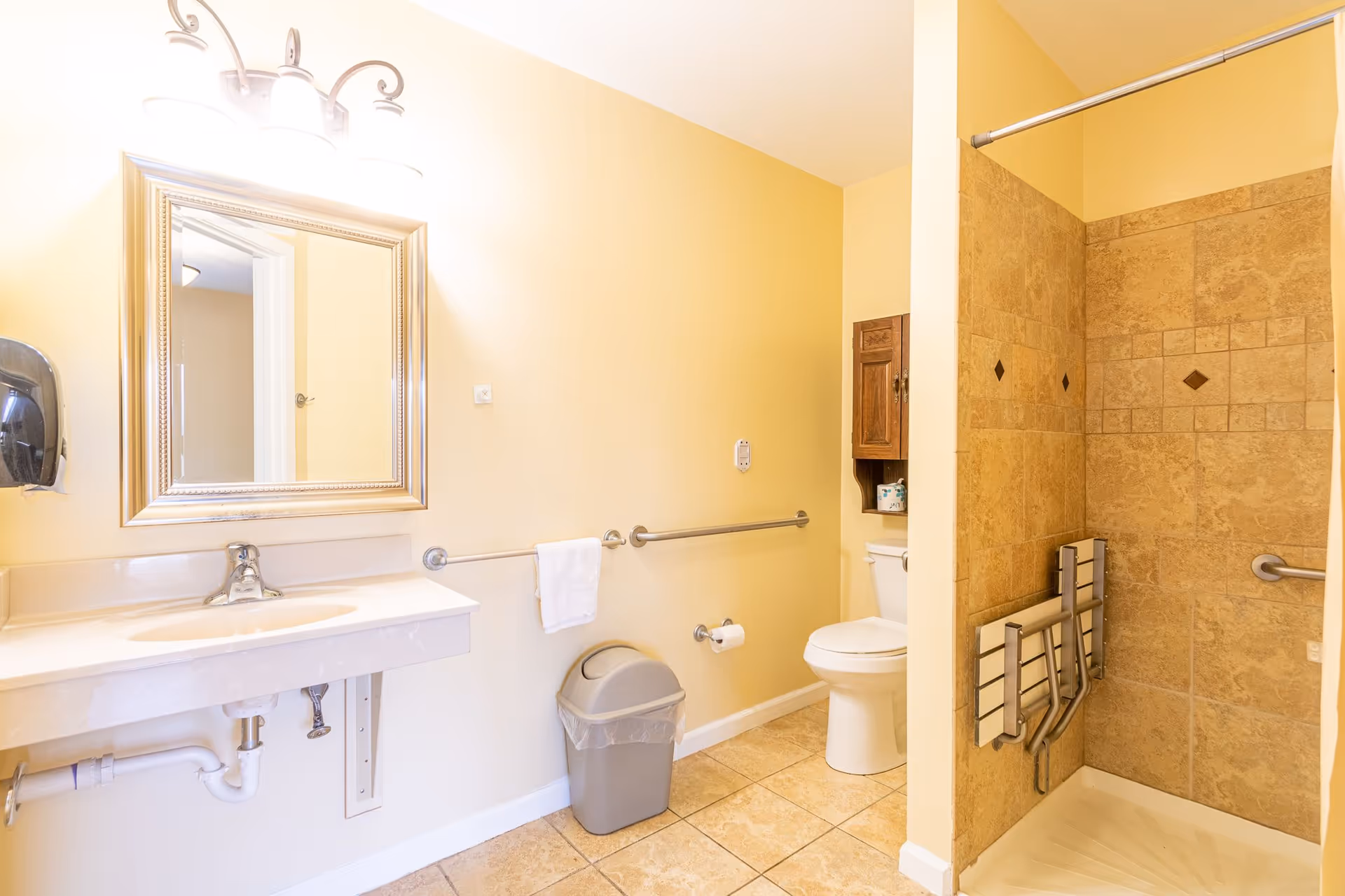 A clean and accessible bathroom with beige tiled floor and walls. The bathroom features a white sink with a large framed mirror above it, a toilet with a wooden cabinet above, a trash can, and a walk-in shower with grab bars and a foldable shower seat. The walls are painted light yellow.
