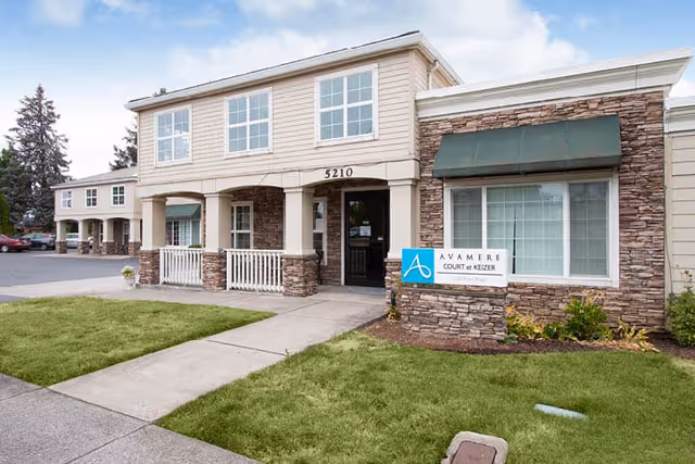 Exterior front view of Avamere Court at Keizer building with stone and beige siding, a green awning over a large window, a sidewalk leading to the entrance, and a sign displaying the facility name.