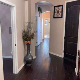 Interior hallway with dark wooden floors and beige walls. A large decorative vase with dried flowers is placed against the left wall near an open doorway. The hallway leads to a bright room with a window, and there is a dark wooden door on the right side. A framed floral artwork hangs on the wall near the door.