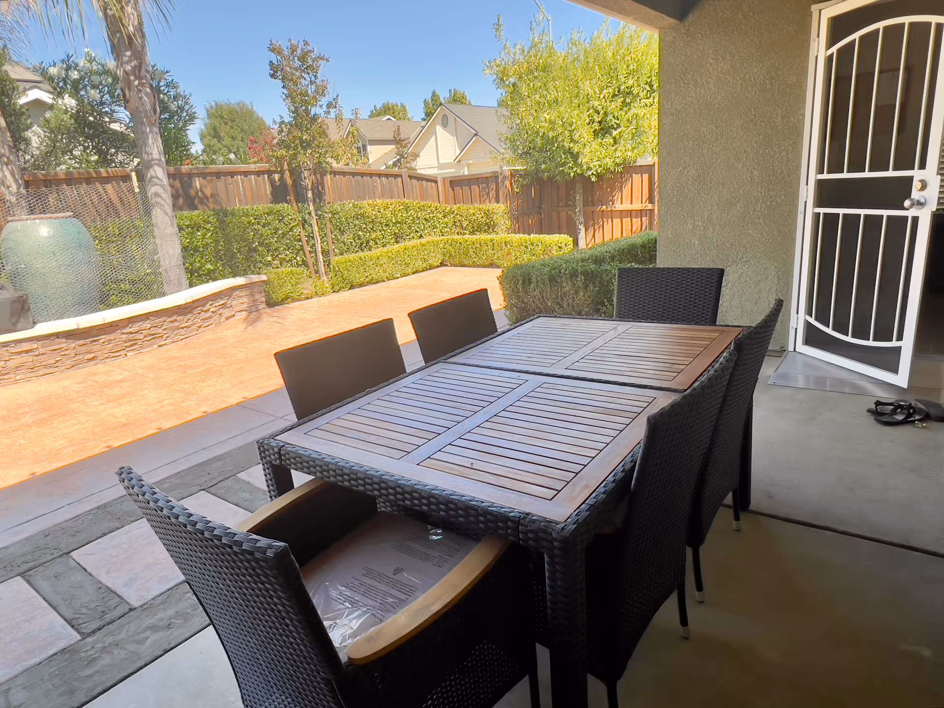 Outdoor patio area with a rectangular wooden table surrounded by six black wicker chairs. The patio is covered and adjacent to a garden with trimmed hedges, trees, and a wooden fence. A white security door is open leading inside the building.