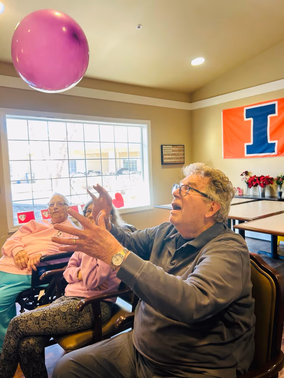 Three elderly individuals sitting in chairs inside a well-lit room. One man in the foreground is reaching up towards a large purple balloon with an enthusiastic expression. Two women sit behind him, one in a wheelchair and the other in a chair, both watching the balloon activity. The room has a large window, a wall decoration with an American flag, and a large orange and blue banner with the letter 'I'.