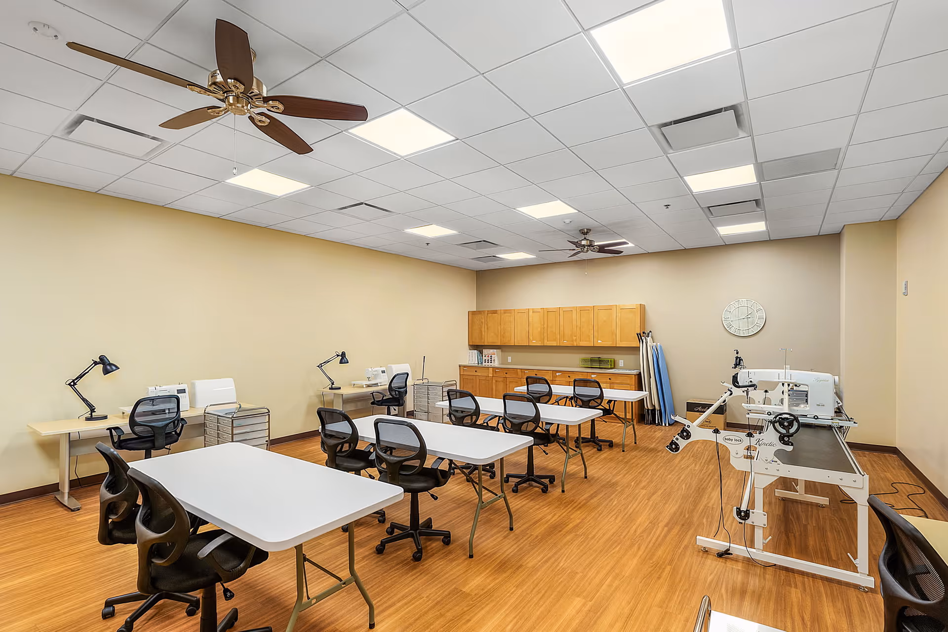 Well-lit multipurpose craft room with folding tables, rolling chairs, sewing machines and wall cabinets.