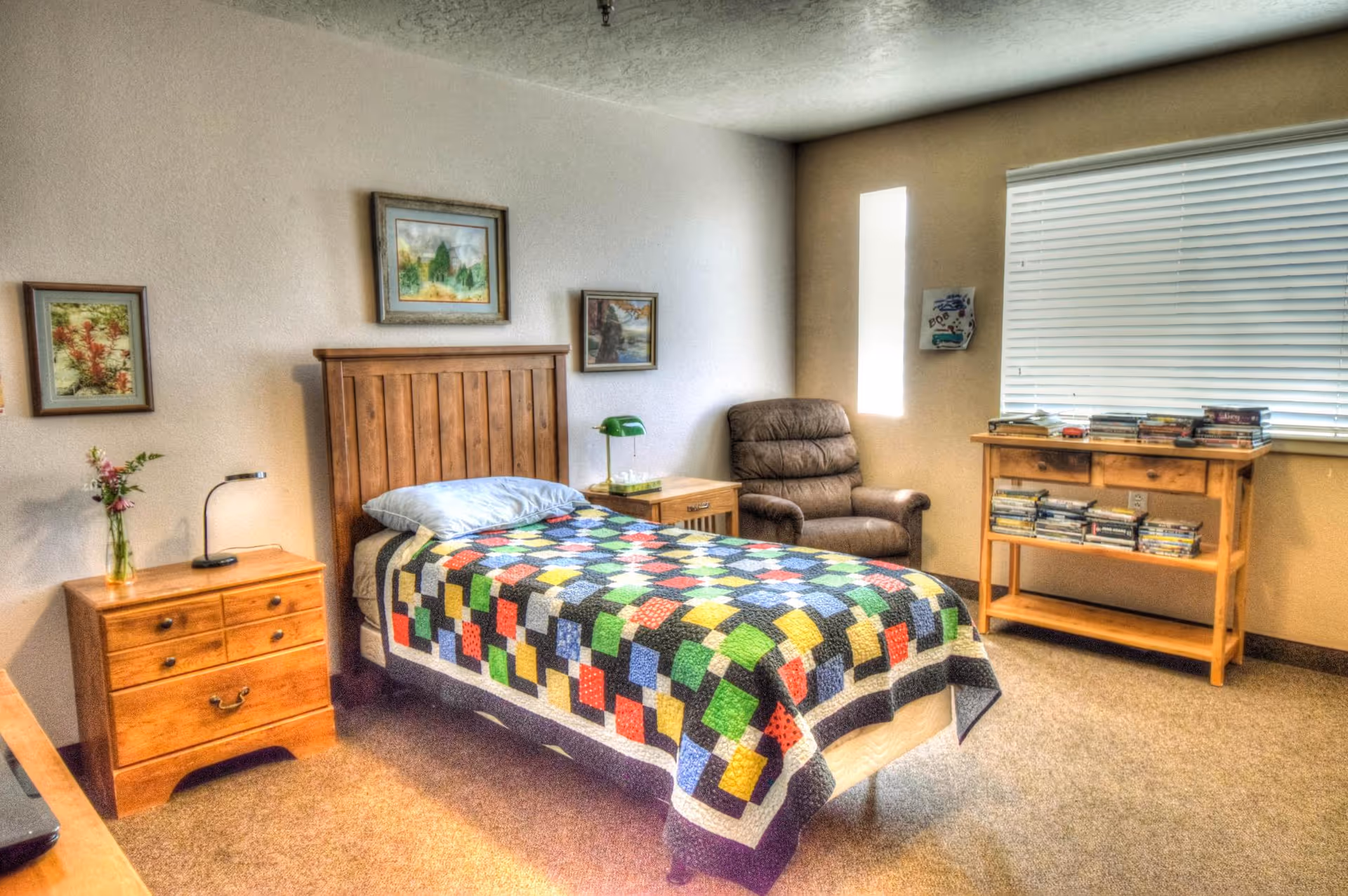A cozy bedroom with a single wooden bed covered with a colorful patchwork quilt. There are two wooden nightstands on either side of the bed, one with a green lamp and the other with a small vase of flowers and a reading lamp. A comfortable brown armchair is placed near a window with closed blinds. A wooden shelf holds a collection of DVDs or books. Three framed paintings hang on the walls.