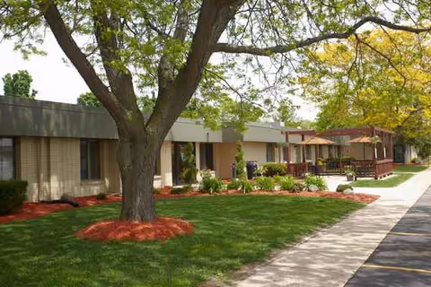 A single-story senior living building with a manicured lawn, trees, and patio seating.