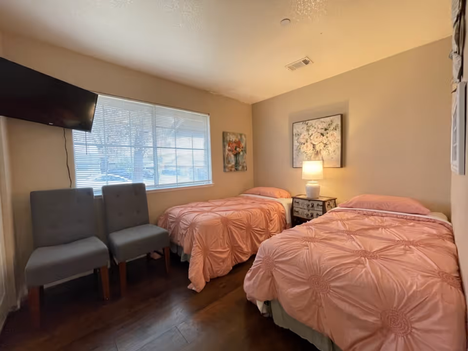 A cozy bedroom with two twin beds covered in matching pink comforters. Between the beds is a small nightstand with a white lamp and a floral painting hanging above it. Two gray chairs are placed near a window with blinds, and a flat-screen TV is mounted on the wall to the left.