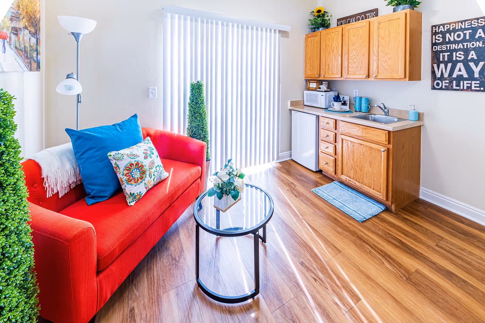A bright and cozy living area featuring a vibrant red sofa with blue and floral cushions, a glass coffee table with a floral arrangement, wooden flooring, and a small kitchenette with wooden cabinets, a sink, a microwave, and a mini fridge. Vertical blinds cover a large window or sliding door, and decorative plants and motivational wall art add warmth to the space.