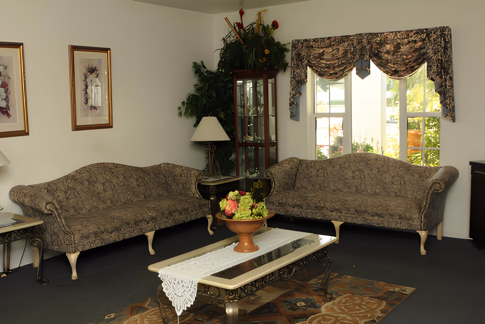 Two patterned sofas facing a glass-topped coffee table with a floral centerpiece in a sunlit sitting room.
