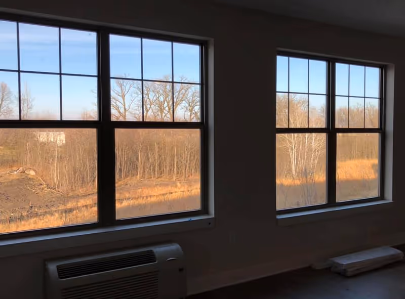 View through two large windows showing a landscape with leafless trees and dry grass under a clear blue sky. Inside, there is a wall-mounted air conditioning unit below the left window and some stacked materials on the floor near the right window.