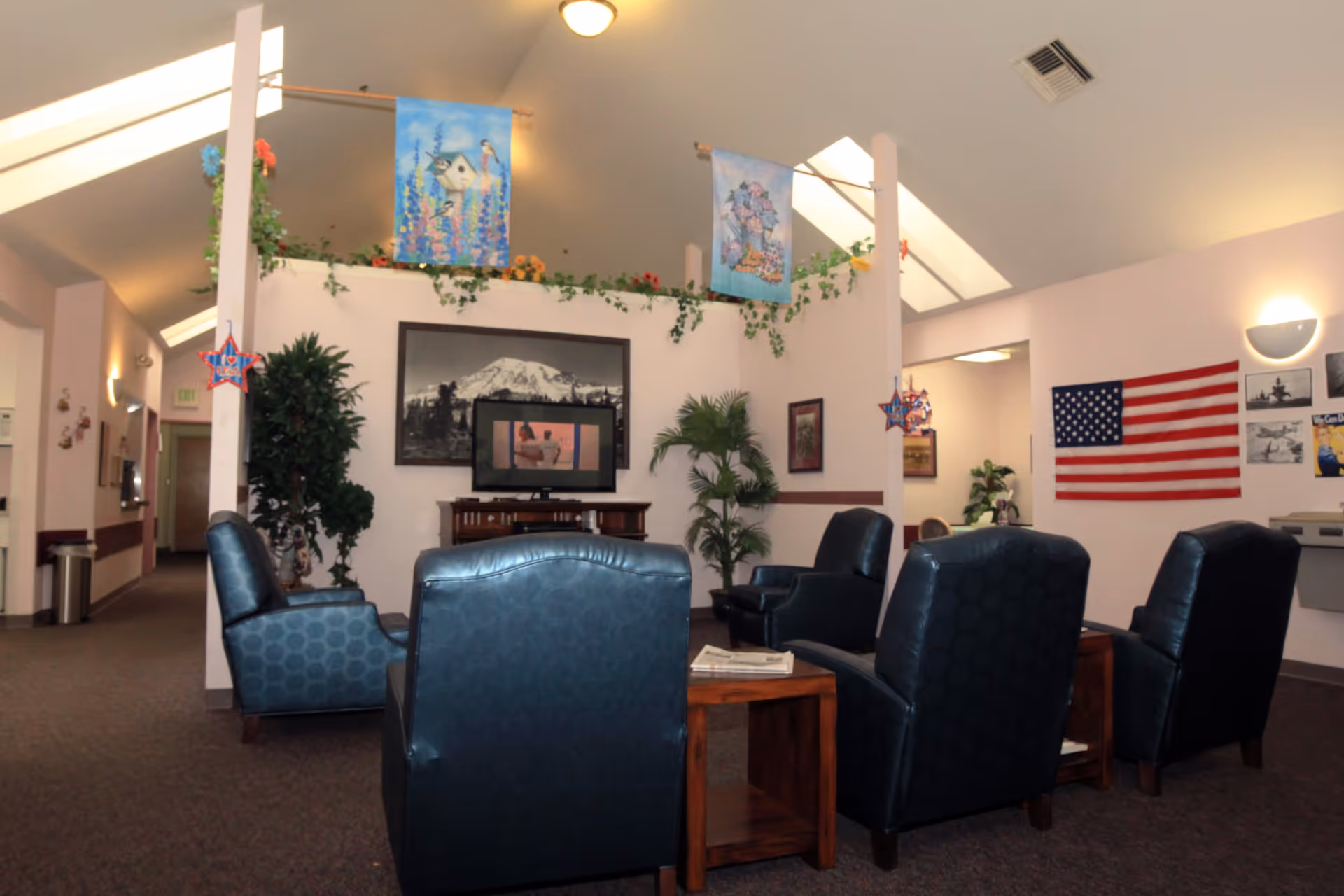 A cozy common area with four dark blue armchairs arranged around a wooden coffee table. The room features a high ceiling with skylights, decorative hanging banners, and green plants. On the wall, there is a large black and white photograph of a mountain, a flat-screen TV, and an American flag with several smaller framed pictures. The space is softly lit with wall sconces and ceiling lights.
