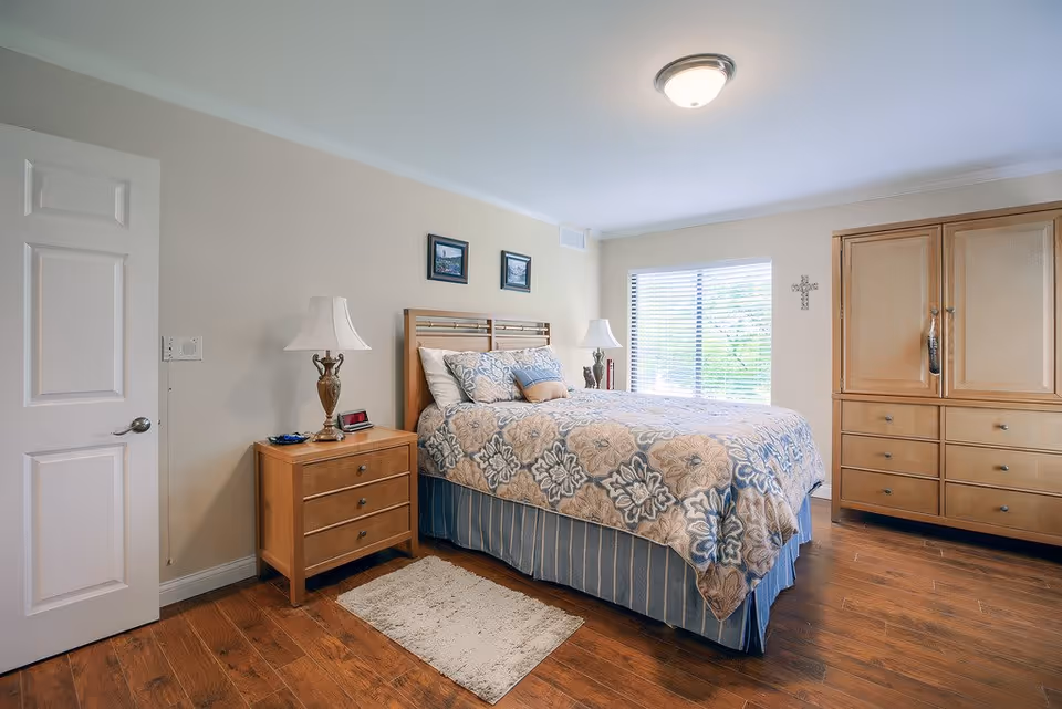 A neatly arranged bedroom with a wooden bed frame and a bed covered with a floral patterned comforter and multiple pillows. There is a wooden nightstand with a lamp and some small items on it to the left of the bed. A window with blinds allows natural light into the room. On the right side, there is a large wooden dresser with multiple drawers. The floor is wooden, and a small rug is placed beside the bed. The walls are light-colored with two small framed pictures above the bed and a decorative cross on the wall near the window.