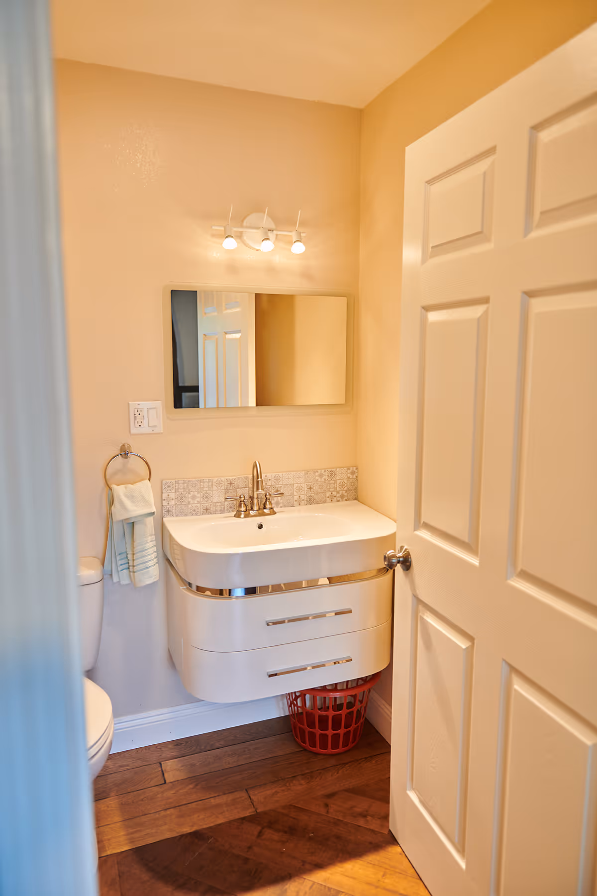 Small bathroom with a white floating sink and cabinet, a mirror above the sink, a towel ring with a hand towel, a toilet partially visible, and a red laundry basket on the wooden floor. The bathroom door is open, showing beige walls and a three-light fixture above the mirror.