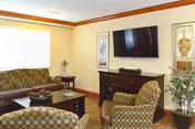 A cozy living room area with patterned armchairs and a sofa around a wooden coffee table. A flat-screen TV is mounted on the wall above a dark wooden cabinet. The room has light-colored walls with crown molding and two framed decorative art pieces on either side of the TV. A large window with blinds lets in natural light, and there is a potted plant in the corner.