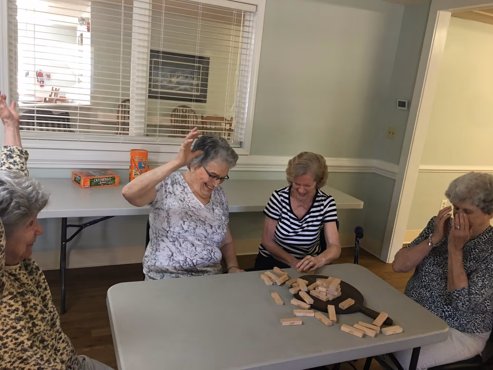 Four elderly women sitting around a table playing a game with wooden blocks. They appear to be enjoying themselves, with one woman raising her hand and another covering her face in amusement. The room has light-colored walls and a window with blinds looking into another room with chairs and a table.