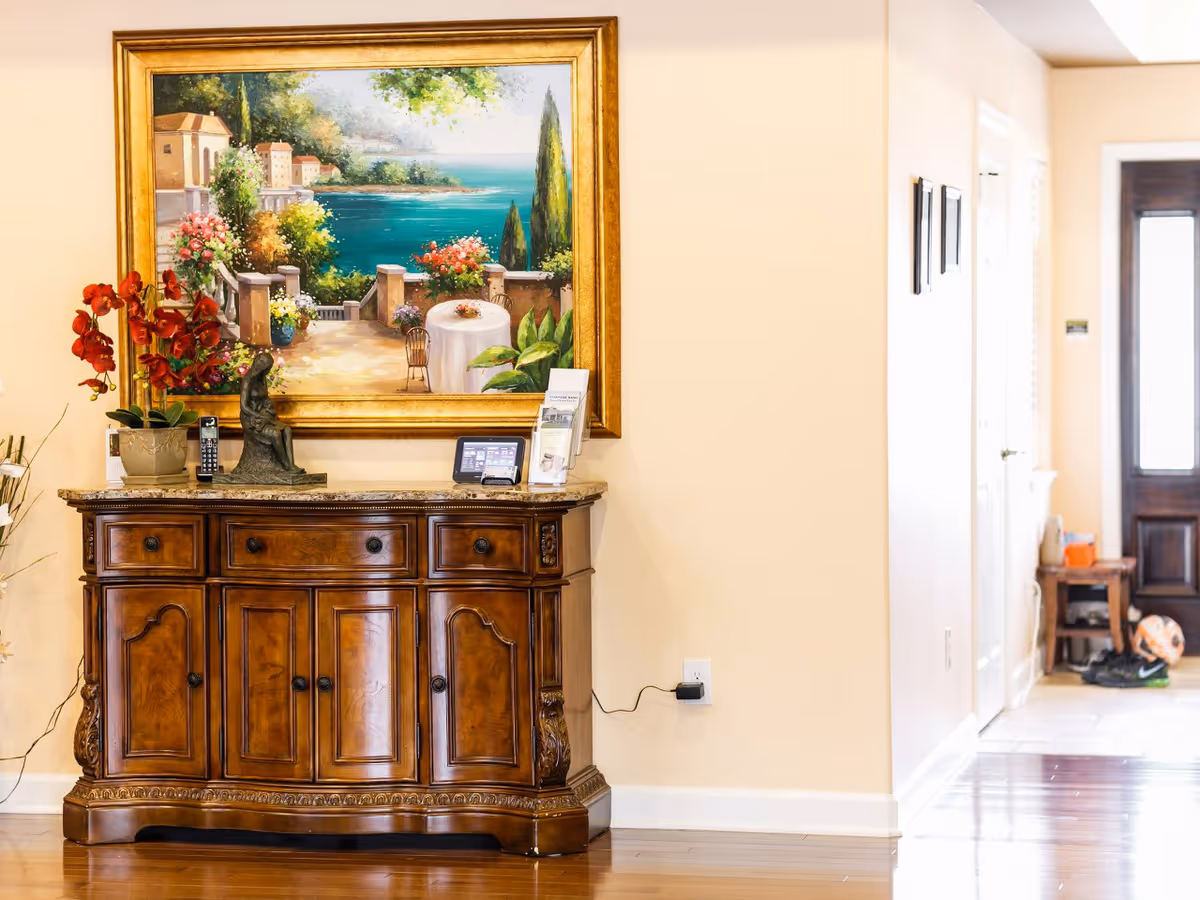 A wooden sideboard with ornate detailing stands against a beige wall. On top of the sideboard are a potted red orchid, a small statue, a cordless phone, and a digital photo frame. Above the sideboard hangs a large framed painting depicting a scenic coastal view with a terrace, flowers, and the ocean. To the right, there is a hallway leading to a door with a window, and some shoes and a ball are visible on the floor.