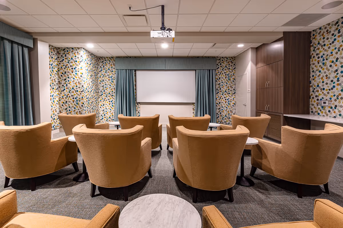A small screening room with rows of tan armchairs facing a white projection screen, patterned walls, and teal curtains.