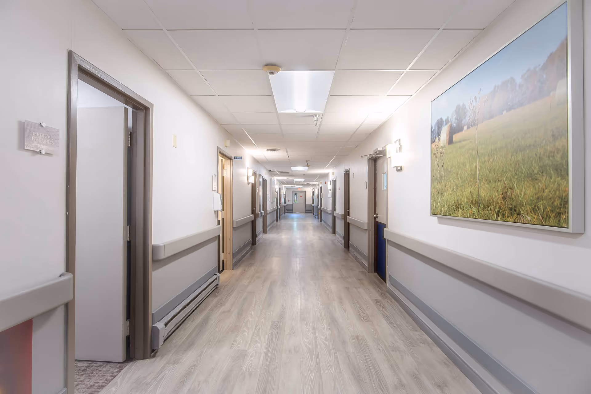 A long, well-lit hallway in a senior living facility with light-colored walls and wood-patterned flooring. Several closed doors line both sides of the corridor, and a large framed picture of a grassy field hangs on the right wall. Handrails run along both sides of the hallway for support.