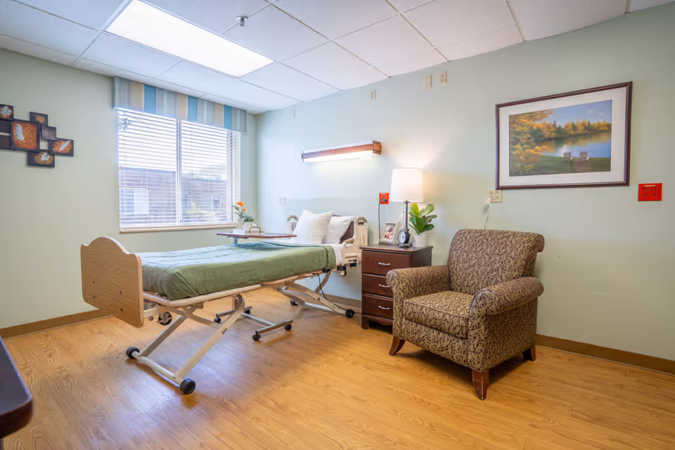 A senior living bedroom with an adjustable bed, wooden nightstand, upholstered armchair, and a window letting in natural light.
