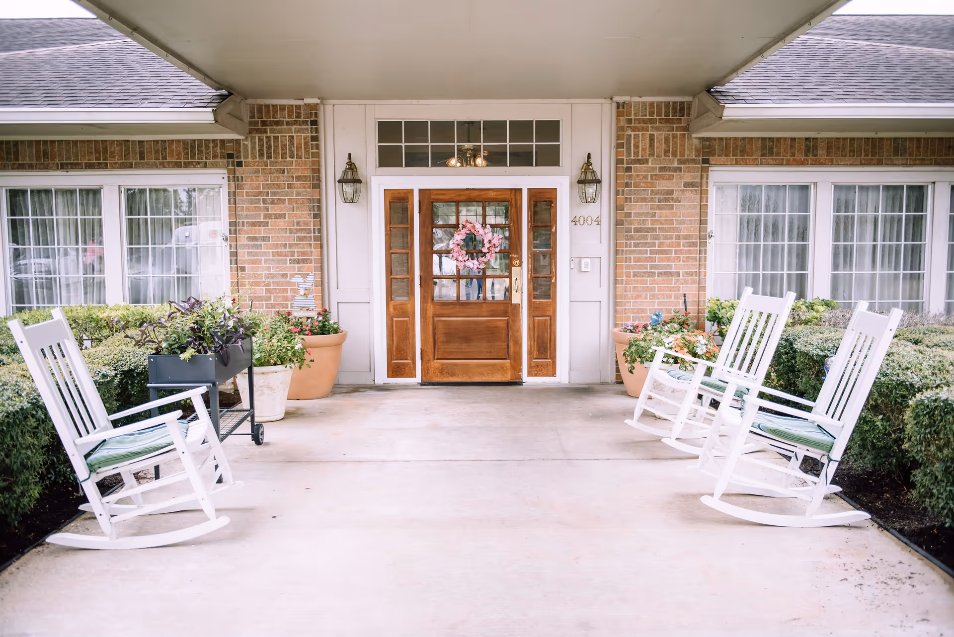 Covered entrance area of a building with a wooden door decorated with a pink floral wreath, flanked by two wall-mounted lantern lights. There are large windows with white curtains on either side of the door. Four white rocking chairs with green cushions are placed on the concrete floor, two on each side. Large potted plants and bushes surround the entrance.