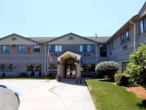 Front exterior view of a two-story senior living facility building named Woodridge, with a covered entrance, American flag, and well-maintained lawn and shrubs under a clear blue sky.