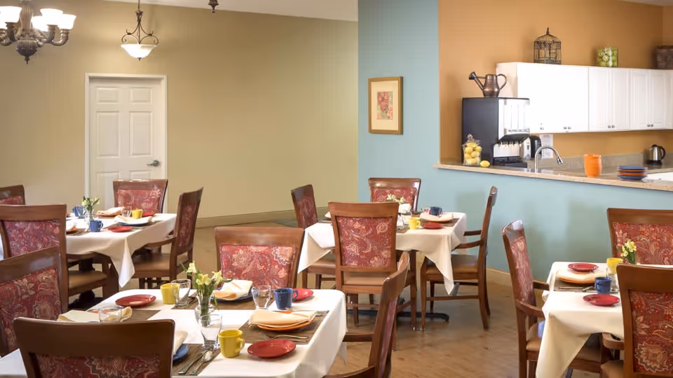 A dining room with several tables covered in white tablecloths, each set with colorful plates, cups, napkins, and silverware. The chairs have wooden frames with red patterned upholstery. The room has a light yellow wall on one side and a blue wall with a counter and white cabinets on the other. There are decorative items on the counter and a framed picture on the wall.