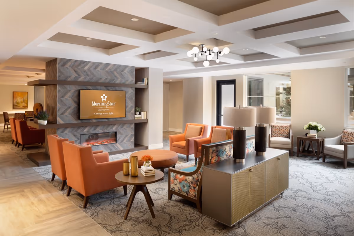 A modern lobby interior featuring orange armchairs, a decorative table with vases, a television mounted on a textured wall, and stylish lighting fixtures.