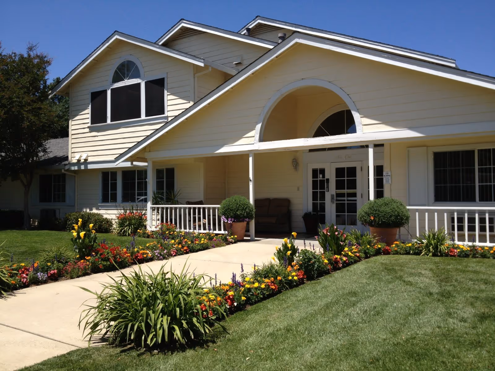 Front exterior view of a light yellow senior living facility building with a well-maintained garden featuring colorful flowers and green shrubs along a concrete walkway leading to a covered porch entrance with seating.