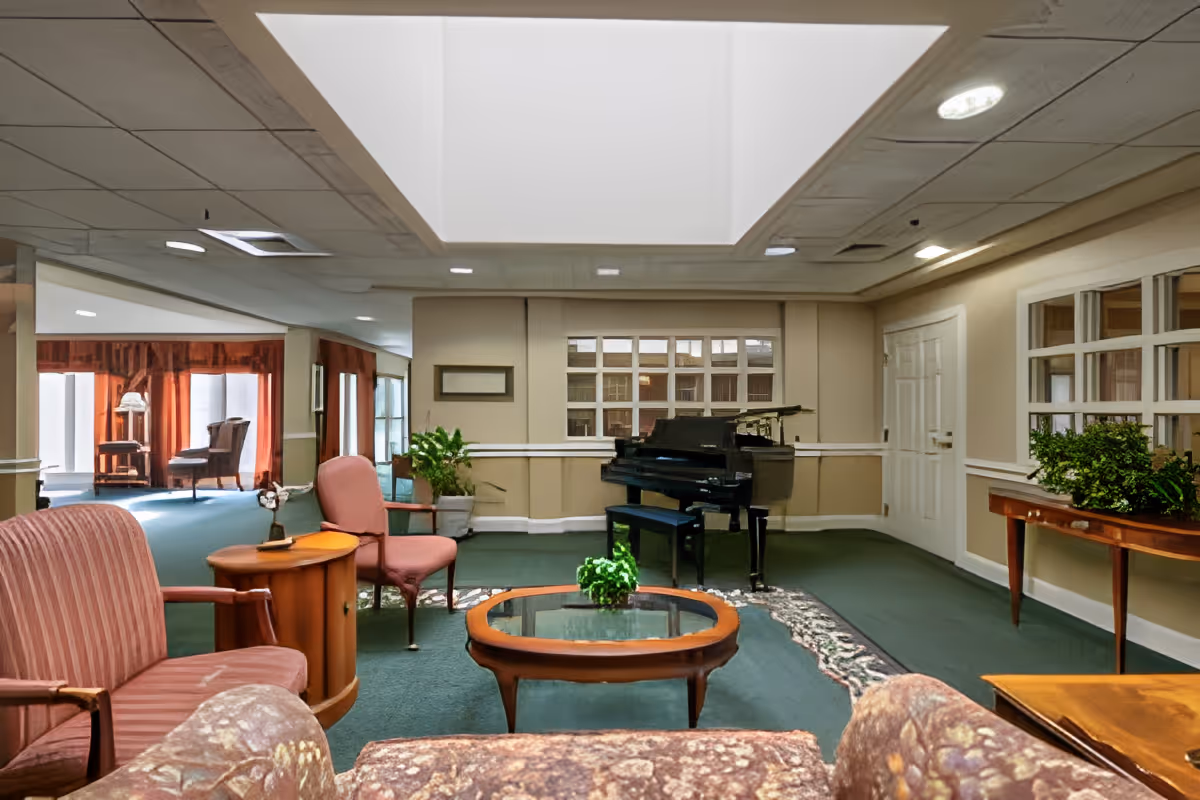 Senior living common lounge with upholstered seating, coffee tables, plants, and a black grand piano beneath a large skylight.