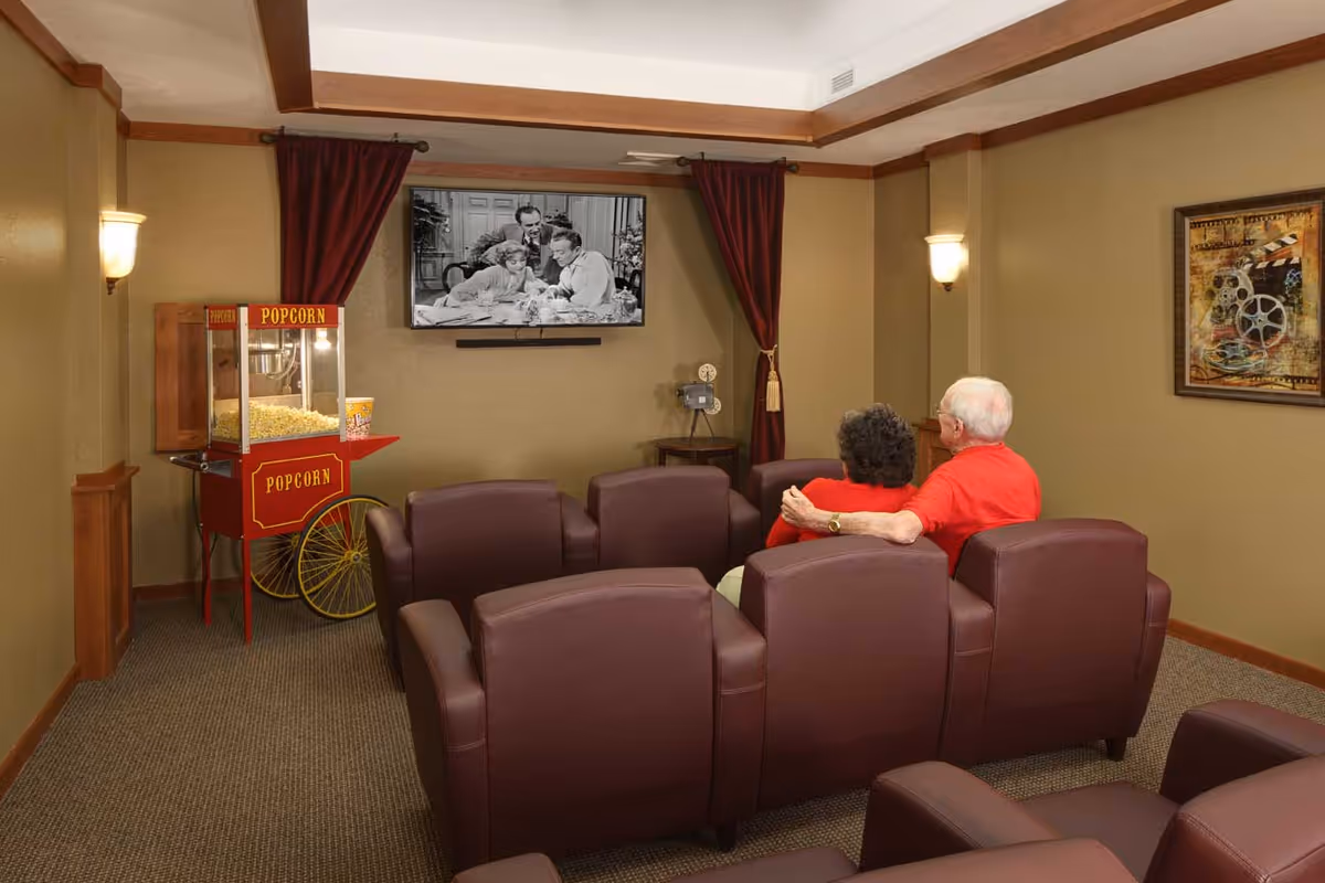 Two elderly people sit in maroon reclining seats in a small theater-style room watching a black-and-white movie, with a popcorn machine to the side.