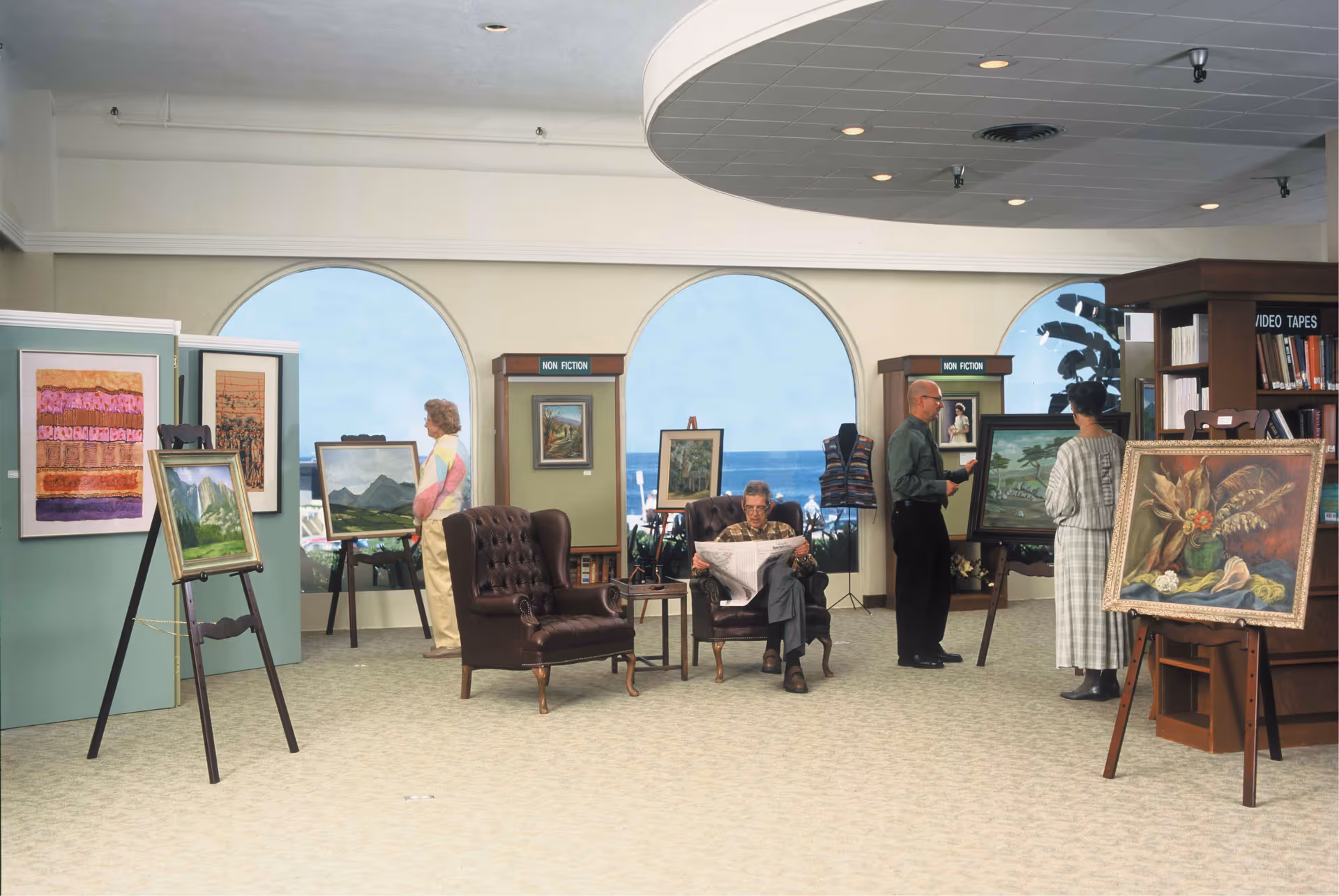 An interior room with large arched windows showing an ocean view. Several paintings are displayed on easels and walls. Three people are observing the artwork, and one person is seated in a leather armchair reading a newspaper. Bookshelves labeled 'NON FICTION' and 'VIDEO TAPES' are visible in the background.