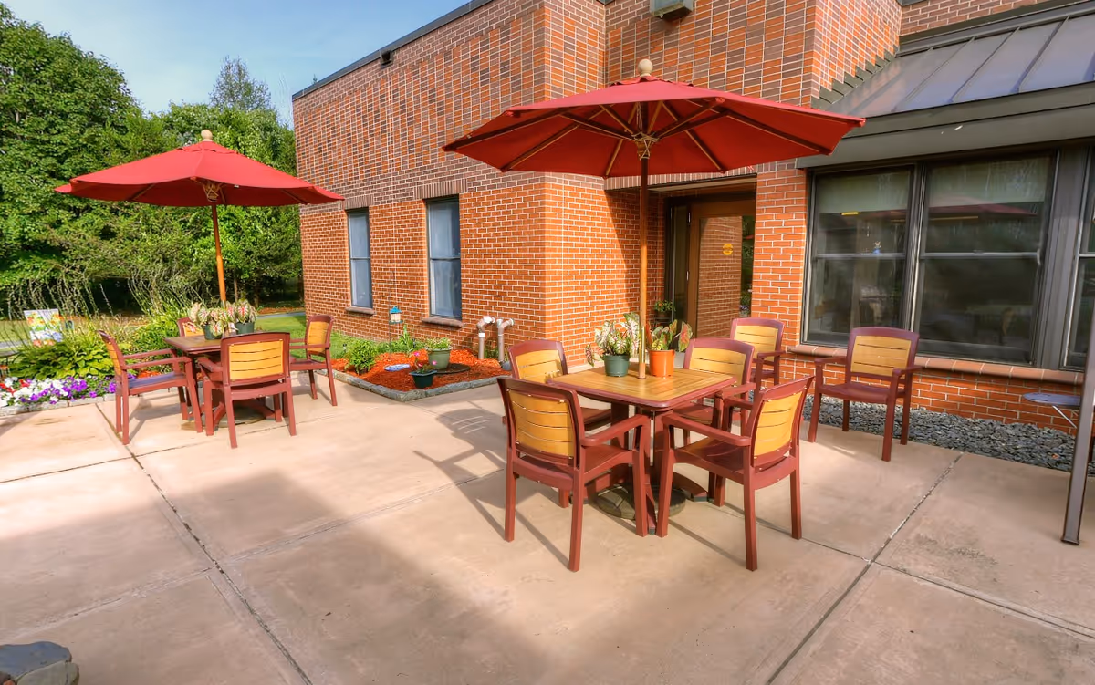 Outdoor patio area with two square wooden tables each surrounded by four chairs with wooden backs and red frames. Each table has a large red umbrella providing shade and small potted plants as decoration. The patio is adjacent to a brick building with windows and a door. There are green trees and plants in the background.