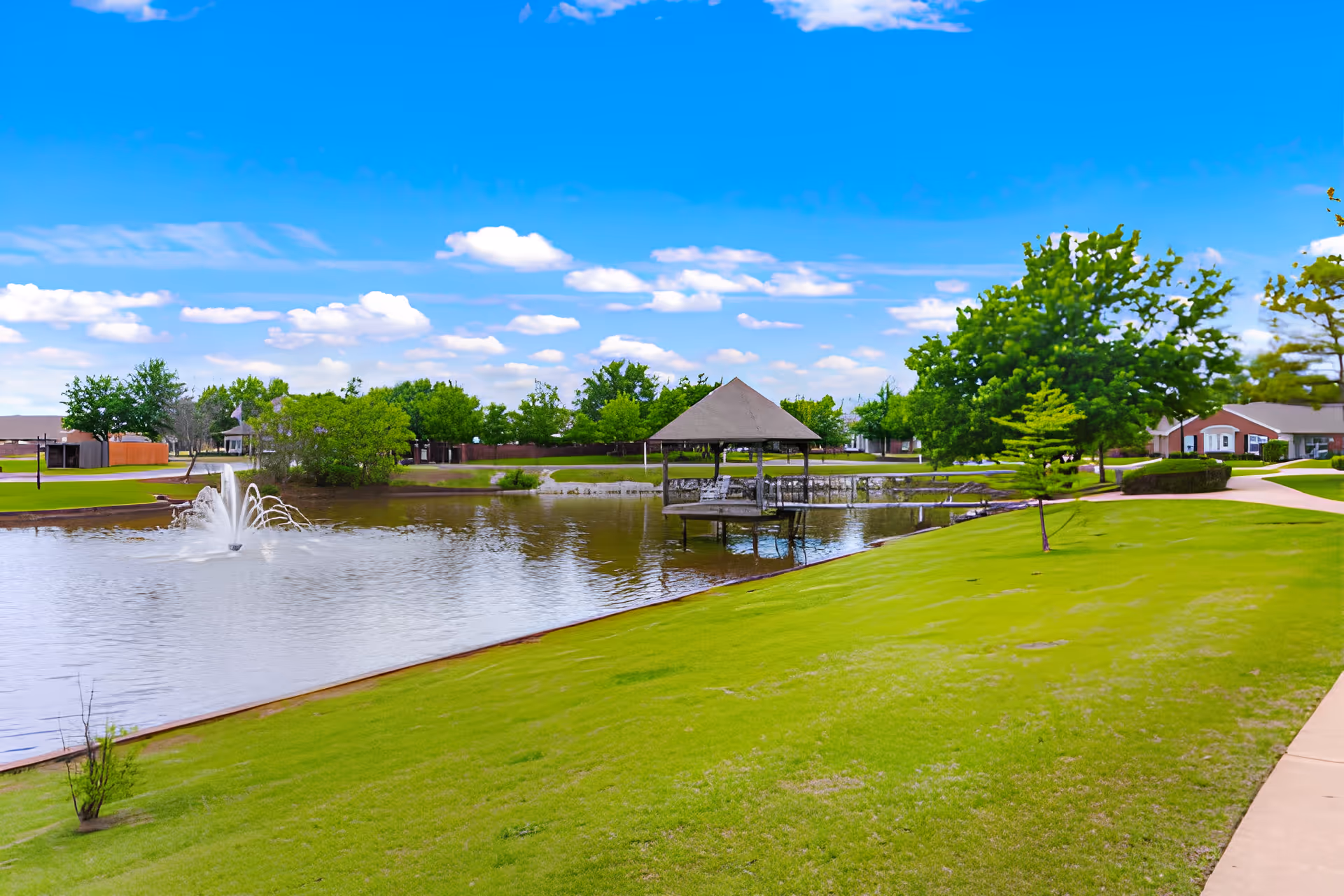 A scenic outdoor area at Elison Independent Living of Statesman Club featuring a small lake with a water fountain, a gazebo on stilts over the water, lush green grass, trees, and a clear blue sky with scattered clouds.