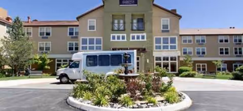 Three-story beige senior living building with a circular landscaped island, fountain, and white shuttle.