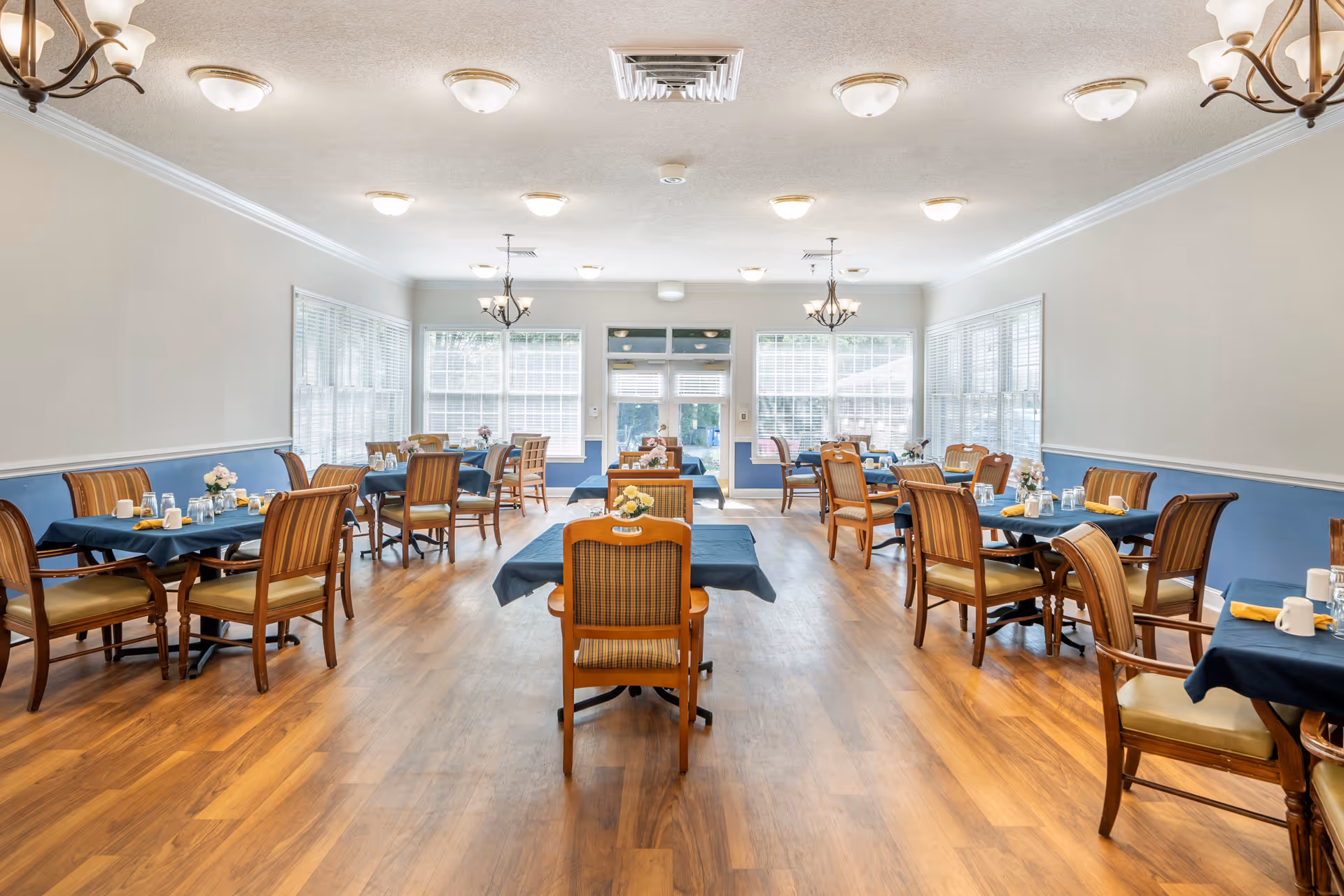 A bright dining room with multiple wooden tables covered with dark blue tablecloths, each set with glasses, cups, and yellow napkins. The room has wooden chairs with cushioned seats and backs, large windows with white blinds letting in natural light, and chandeliers and ceiling lights providing additional illumination. The floor is wooden, and the walls are painted light with a blue wainscoting.