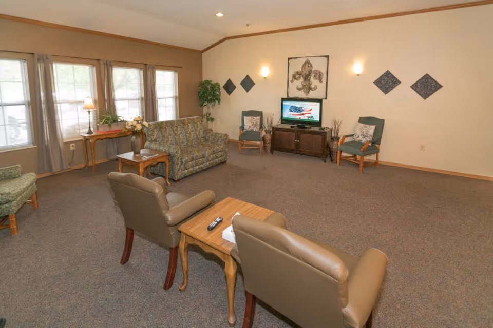 A senior living facility common area with a patterned sofa, two green armchairs, two brown leather chairs, a wooden coffee table with a remote and tissue box, a TV on a wooden stand displaying an American flag, wall decorations including a fleur-de-lis artwork, and large windows with curtains letting in natural light.